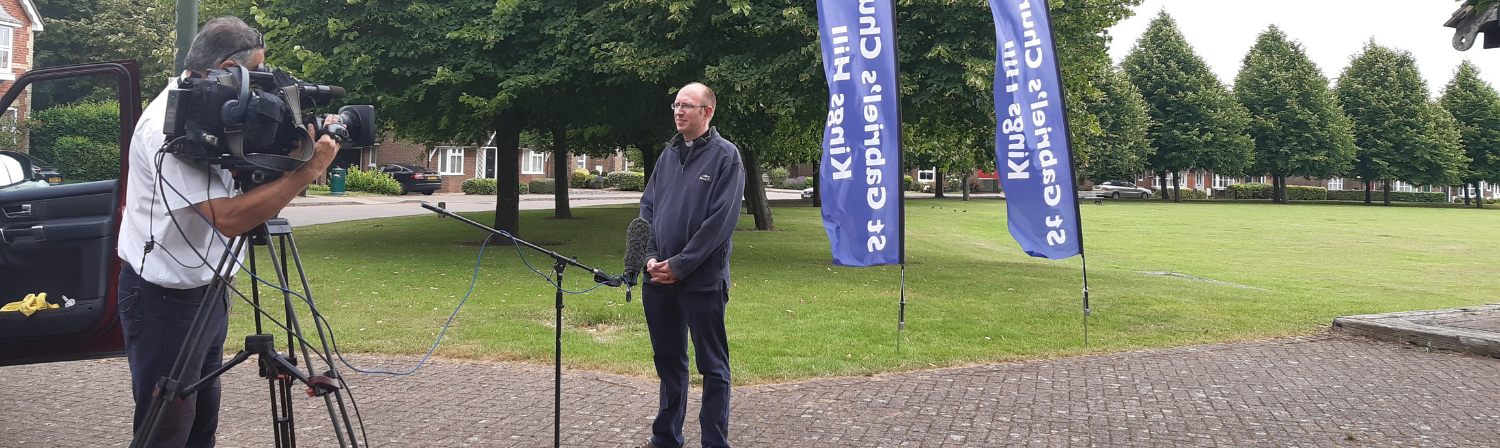 A vicar stands ready to give an interview to a journalist with a video camera.