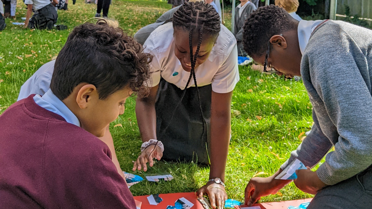 Four children kneel on the grass as they arrange pieces of paper on a red piece of cardd