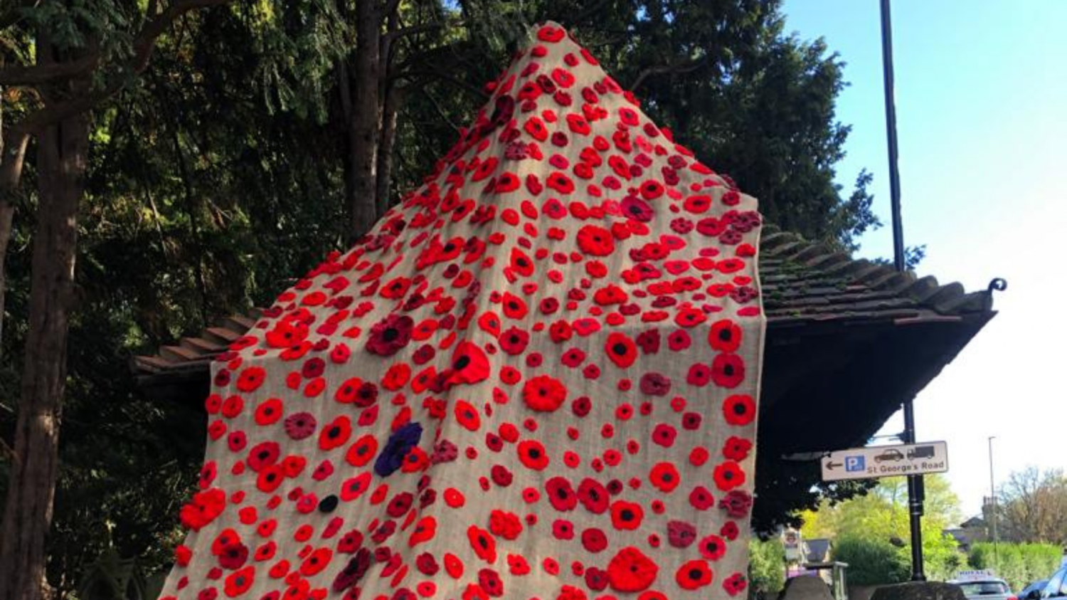 Bright red knitted and crocheted poppies of different sizes hand on a brown sackcloth draped from a tree over the gated entrance into the church yard. 