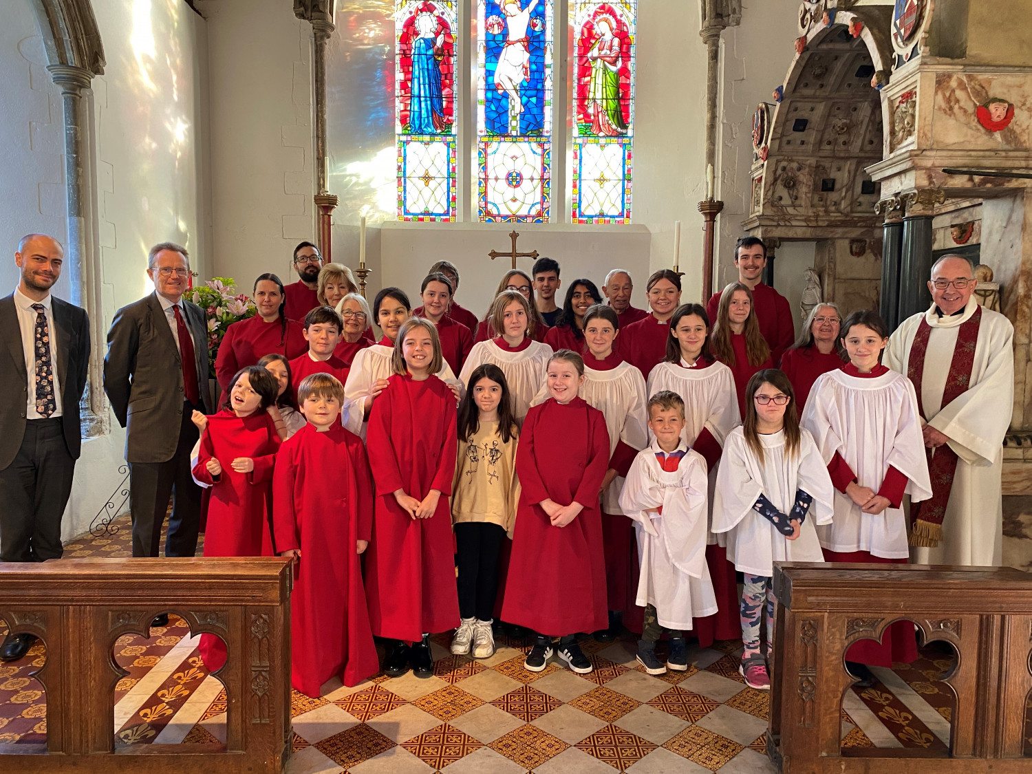 A mixed choir of robed children, young people, and grown-ups stand beneath a stained glass window in the the church and smile.