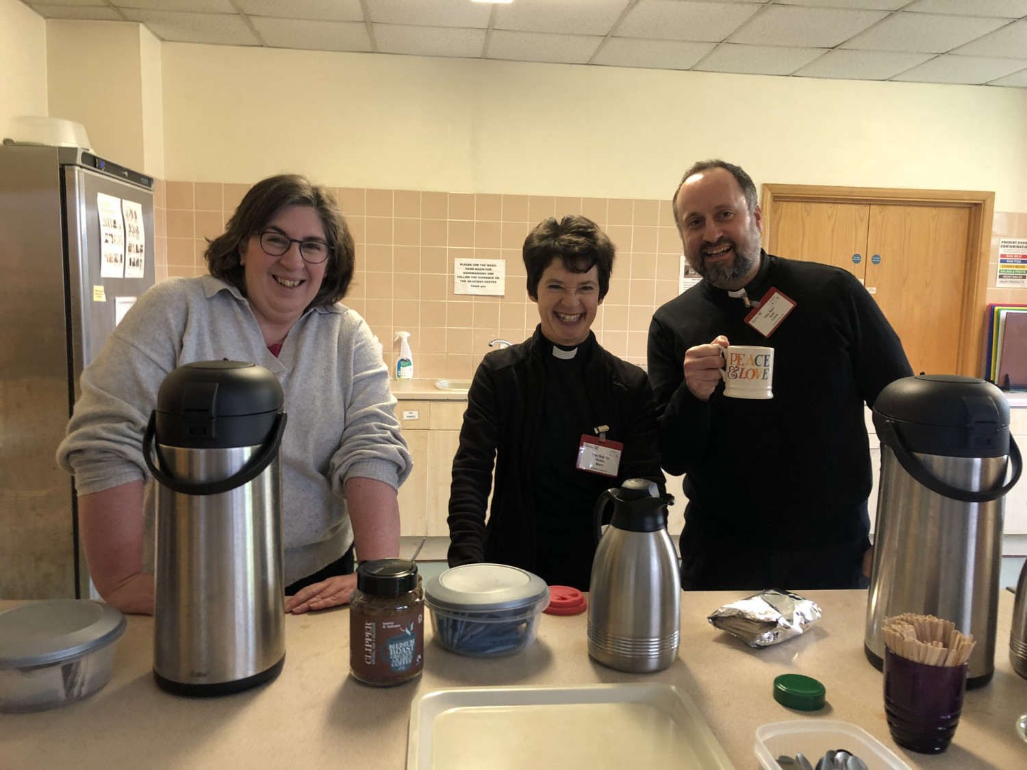 Standing behind a serving hatch, three members of Synod smile as the prepare to distribute tea and coffee from urns.